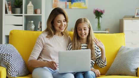 Cheerful-good-looking-mother-and-daughter-sitting-together-on-the-yellow-couch-and-having-a-videochat-on-the-laptop-computer-in-the-cozy-living-room.-Indoor.